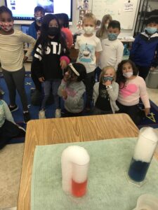 First graders watch a chemical reaction of colored water bubbling over onto the table.