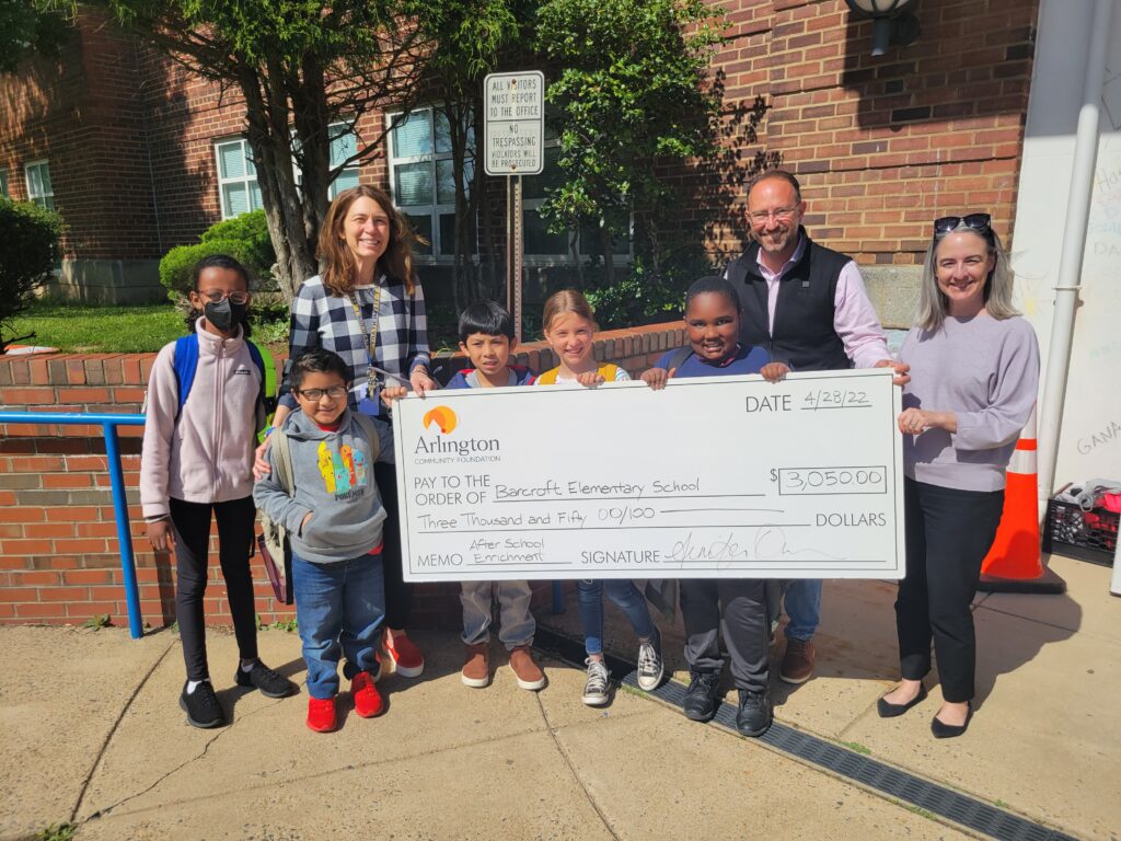 El director y los estudiantes de la Escuela Primaria Barcroft sonriendo y recibiendo un gran cheque para una donación fuera de Barcroft.